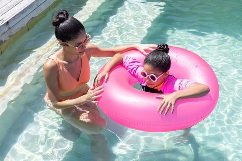 African american mother supervising daughter on bright pink float in sunny backyard pool