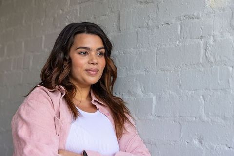 Confident woman in pink overshirt showing smartwatch, crossed arms, gray brick backdrop