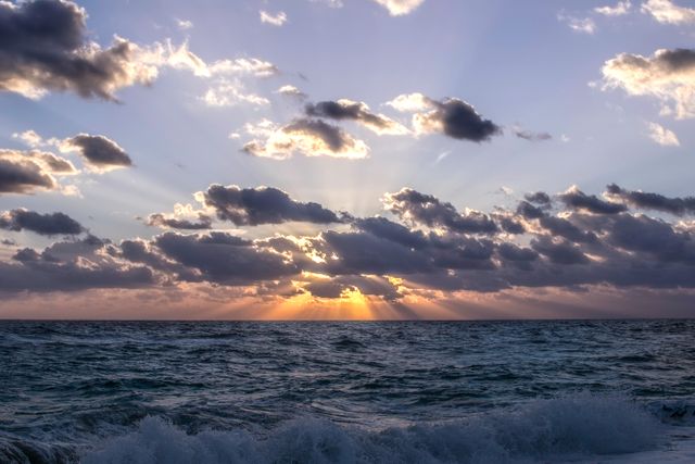 This image captures a dramatic sunset over the ocean, with rays of sunlight breaking through clouds and waves crashing. Ideal for use in travel brochures, motivational posters, or as a background for websites highlighting natural beauty.