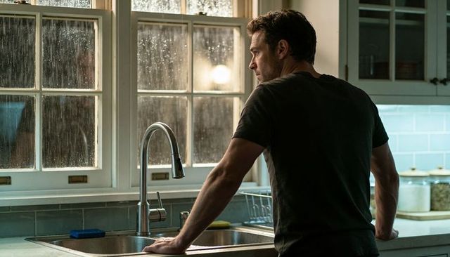 Man leaning at kitchen sink gazing through rainy window in evening light