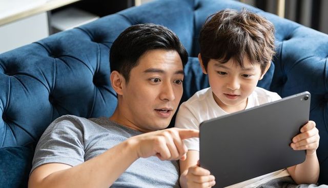 Father and son sharing tablet on blue sofa showing digital learning and family bonding