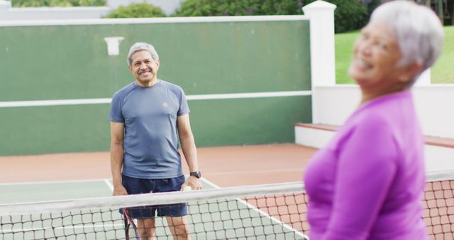 Senior couple is pictured enjoying a game of tennis on an outdoor court. The man is standing at the net in athletic wear, smiling and looking relaxed, while the woman in the foreground wearing a purple shirt also appears joyful. This scene highlights the importance of staying active and enjoying recreational activities during older age. Ideal for content related to senior health, fitness, active lifestyles, and recreational activities.