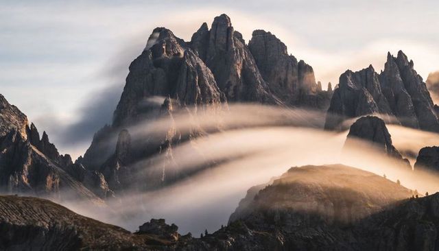 Jagged alpine peaks rising through bands of rolling fog over sunlit rocky ridge