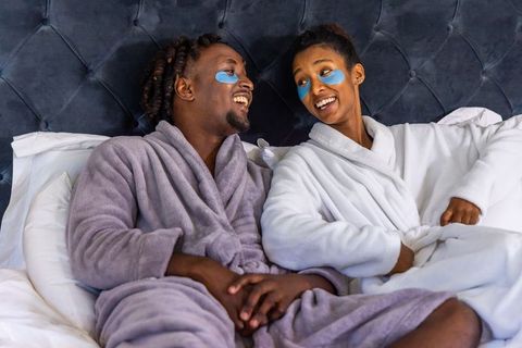 African american couple relaxing in bed wearing eye patches and plush robes, laughing