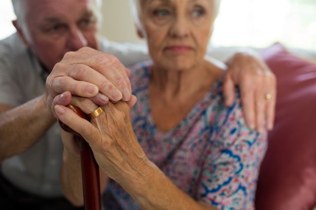Senior couple sitting together at home, holding hands with a walking stick, showing support and companionship. Ideal for use in articles or advertisements about elderly care, aging, retirement, and family support.