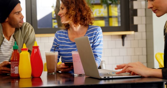 Group of friends talking and enjoying smoothies while seated at a coffee shop. One person is working on a laptop. The scene captures a casual, relaxed setting perfect for highlighting social interaction, modern lifestyle, or promoting cafes and collaborative workspaces.