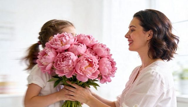 Mother and daughter exchanging large pink peony bouquet in bright kitchen, smiling together