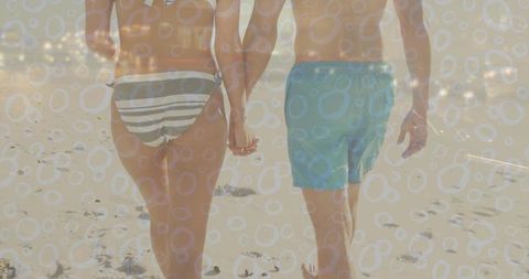 Couple walking hand-in-hand on sunlit beach with striped bikini bottoms and aqua trunks