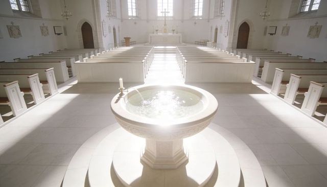 Sunlit baptismal font in white church interior with altar cross and symmetrical pews