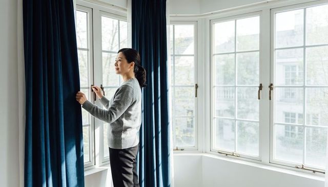 Asian woman opening navy curtains at bay window, sunlit minimalist room, looking outside