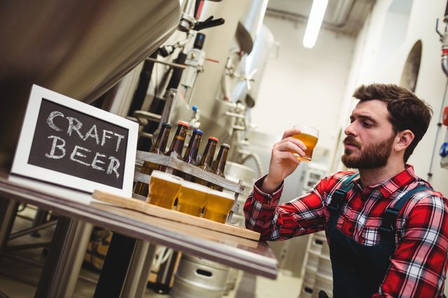 Young brewer in plaid shirt and apron examining a glass of craft beer in an industrial brewery. Chalkboard sign with 'Craft Beer' written on it and several beer bottles on a table. Ideal for use in articles about craft beer production, brewery tours, beer tasting events, and the craft beer industry.