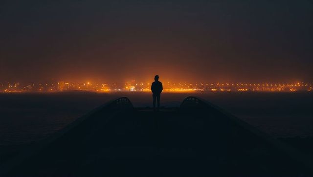 Solitary Figure on Waterfront Pier at Night Overlooking City Skyline