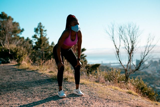 Fit african american woman wearing face mask, exercising in countryside. healthy active lifestyle and outdoor fitness during coronavirus covid 19 pandemic.