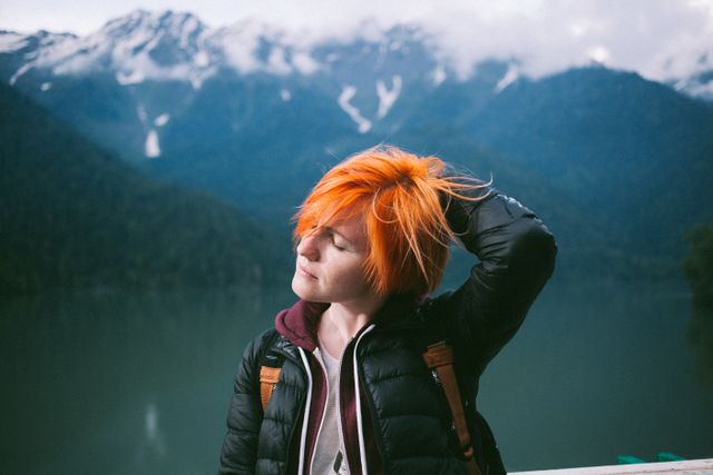 Young red-haired woman enjoying serene mountain lake view, surrounded by misty mountains; perfect for travel blogs, adventure journals, outdoor gear advertisements, and wellness content emphasizing nature's tranquility and beauty.