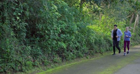 Diverse couple jogging on mossy forest trail wearing sportswear and holding phone