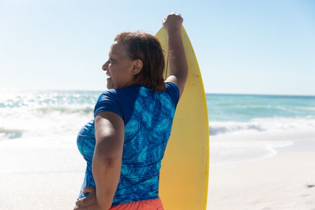 Rear view of senior african american woman standing with hand on hip holding surfboard at beach. unaltered, active lifestyle, aquatic sport and holiday concept.