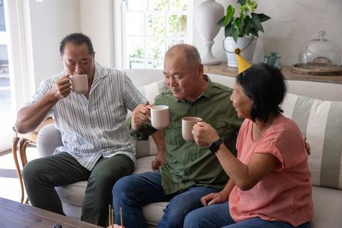 Asian family celebrating with coffee mugs on couch, senior birthday gathering at home