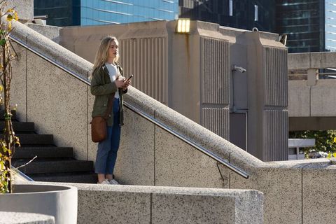 Young woman standing on concrete stairs holding smartphone and coffee, urban lifestyle