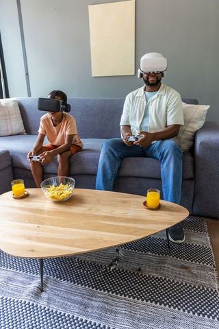 African american father and son playing vr games on couch, bonding with controllers