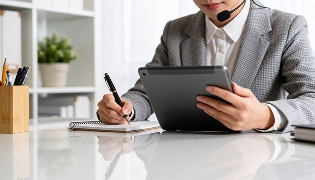 Asian businesswoman holding tablet and taking notes wearing headset in modern office