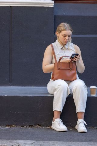Young woman sitting on urban ledge checking smartphone with leather handbag and coffee