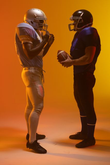 Two male American football players stand face-to-face, creating a sense of competition and intensity. The orange lighting adds a dramatic effect, highlighting the athletes' determination and readiness. This image is ideal for use in sports-related content, advertisements promoting athletic gear, or articles discussing teamwork and sportsmanship.