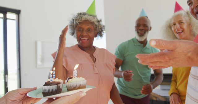 Scene shows a group of senior friends enjoying a birthday celebration with cupcakes and lit candles. They are wearing party hats and engaging in cheerful interactions. Ideal for ads and content highlighting senior living, parties, friendship, aging, and joy in later years.