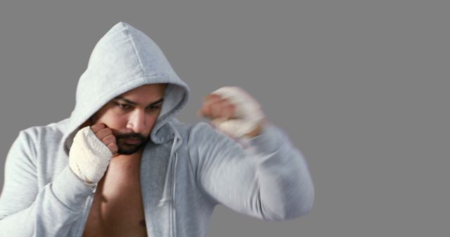 A young, athletic man of diverse ethnicity is captured in a dynamic boxing pose, wearing a hoodie and hand wraps, with copy space. His intense focus and powerful stance convey determination and strength.