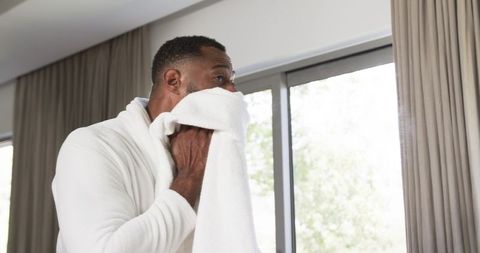 Mature man drying face with towel in bathrobe by sunlit window, relaxed morning ritual