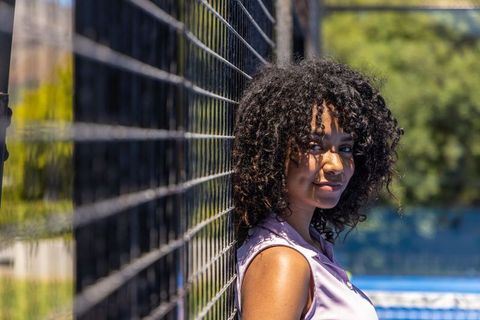 Sunlit african american woman leaning on chain-link fence at blue court, thoughtful portrait