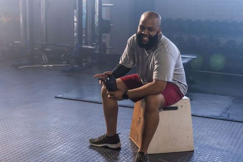 Smiling african man sitting on plyo box holding shaker bottle in moody gym during workout break