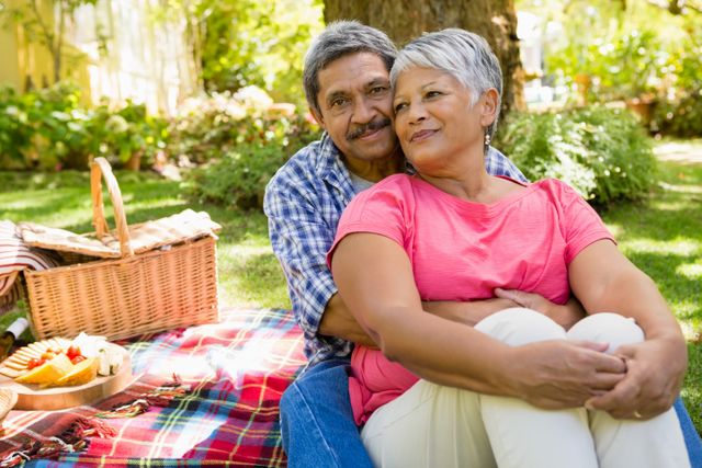 Senior couple sitting on a blanket in a garden, enjoying a sunny day. They are surrounded by greenery and have a picnic basket with food. This image can be used for promoting outdoor activities, retirement lifestyle, senior living, and family bonding.