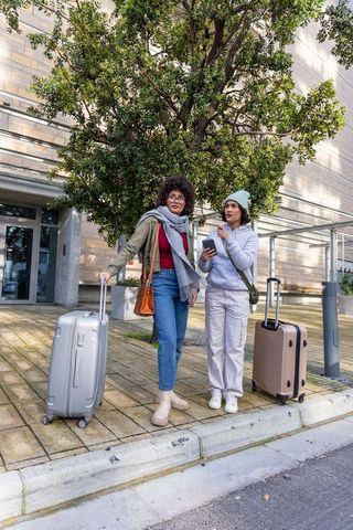 Young women pausing with rolling luggage while checking directions on smartphone