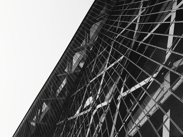 Monochrome image showcasing abstract geometric patterns of a modern architectural structure. Glass building with reflections creating a complex, futuristic design. Ideal for use in architectural design projects, urban themes, visual arts, or as a stylish background for websites and presentations.