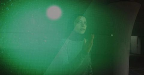 Moody young woman wearing hijab touching neon green digital grid in urban corridor