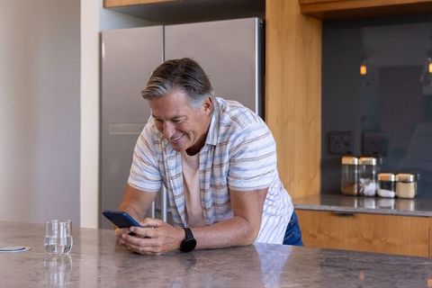 Senior man leaning on kitchen counter using smartphone and smiling in modern home