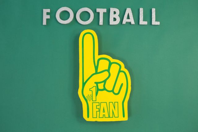 Close-up of a yellow foam hand with 'Football' text on a green background. Ideal for use in sports-related promotions, fan merchandise advertisements, and event marketing materials. Perfect for illustrating enthusiasm and support in sports contexts.