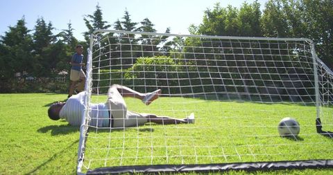 African american man and teen playing backyard soccer, goalie diving, ball in net