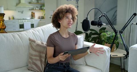 African american woman recording home podcast on sofa with microphone and tablet