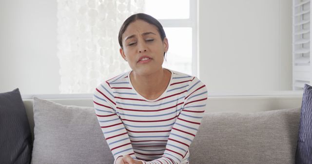 Young woman displaying sadness while sitting on couch in bright living room. She is wearing a striped shirt and appears to be lost in thought or experiencing emotional distress. Suitable for use in articles or blogs about mental health, emotional well-being, or personal stories.