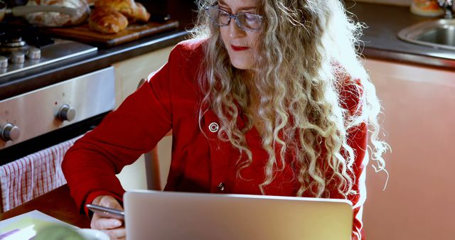 An elderly woman with long silver hair and glasses in a red jacket is focused while working on her laptop in a bright kitchen. A smartphone is held in her right hand. Ovens and baked goods are visible in the background, suggesting a comfortable home environment. Ideal for themes related to elderly technology use, remote work, home comfort, and digital lifestyles.