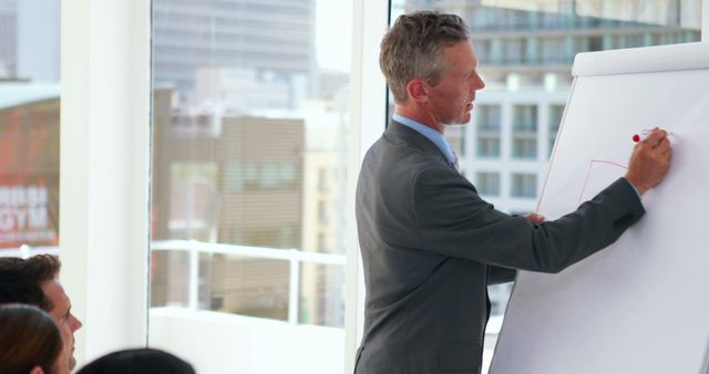 Mid-adult businessman presenting information on whiteboard to colleagues in modern office conference room. Scene suggests teamwork, strategy planning, and leadership in a corporate environment. Useful for articles on business methods, executive training sessions, and corporate teamwork.