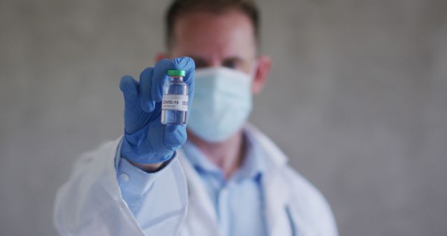 Scientist presenting COVID-19 vaccine vial while dressed in laboratory attire and wearing a mask and gloves. This image can be effectively used in materials promoting vaccine awareness, scientific research, healthcare initiatives, and pandemic-related news.