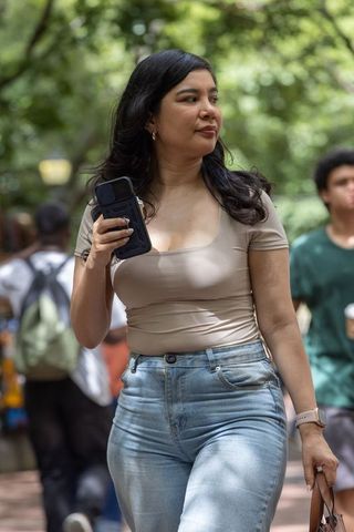 Woman walking on shaded campus path wearing beige top and jeans holding smartphone