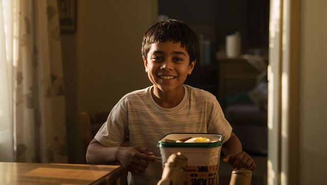 Smiling indian preteen boy holding snack container at sunlit dining table, home lifestyle