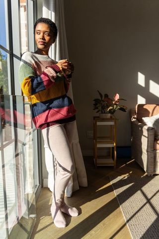 African american woman in chunky striped sweater standing by sunlit sliding door holding mug