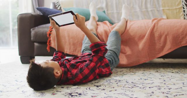 Boy laying on carpet while using tablet. This is great for topics related to childhood, technology use, relaxation at home, or screen time. Can be used in articles about family life, digital devices, kids' activities, or decorating living spaces.