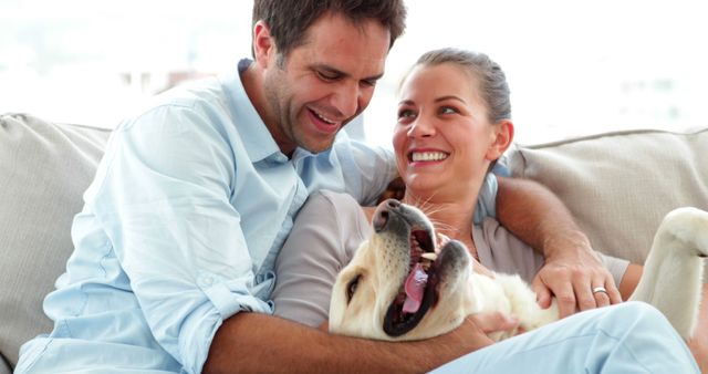 This image shows a joyful couple relaxing comfortably on a couch while playing with their dog. They seem happy and connected, enjoying a peaceful moment at home. This image is perfect for use in advertising for home products, pet care products, or relationship articles, emphasizing joy, bonding, and affection.