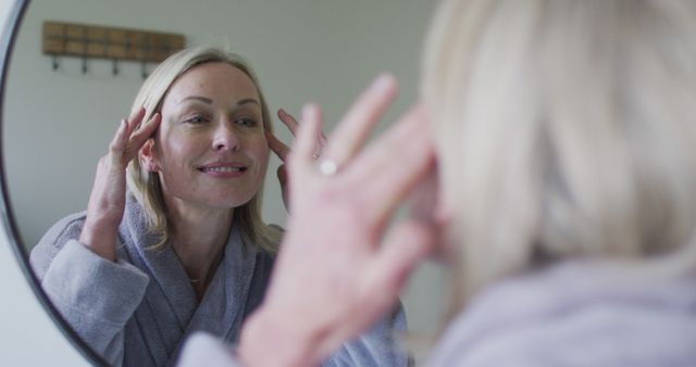 Mature woman smiling at her reflection while applying skincare in bathroom. Useful for topics on beauty routines, wellness, healthy lifestyle, and self-care promotion.