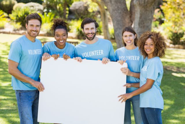 Group of diverse volunteers standing together in a park, holding a blank sign. They are smiling and wearing blue shirts with 'Volunteer' written on them. This image can be used for promoting community events, charity work, social activism, and teamwork initiatives.